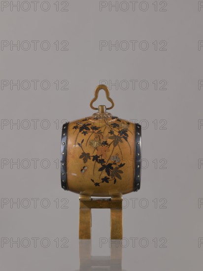 Okimono in the shape of a taiko drum by Nogawa, ca. 1890–1900 Meiji period, 1868–1912. Drum-shaped incense burner, Japanese, Kyoto., Japanese Meiji-period okimono with butterflies, insects, plant motifs, and paulownia decoration. Dimensions: height 15.0 cm; diameter 7.8 cm., An ornate cylindrical object in golden brown rests on a matching stand, decorated with floral motifs in dark and muted colors; it has a curved handle on top in a similar golden tone and dark vertical bands with small round accents on its sides, all set against a plain neutral gray background.