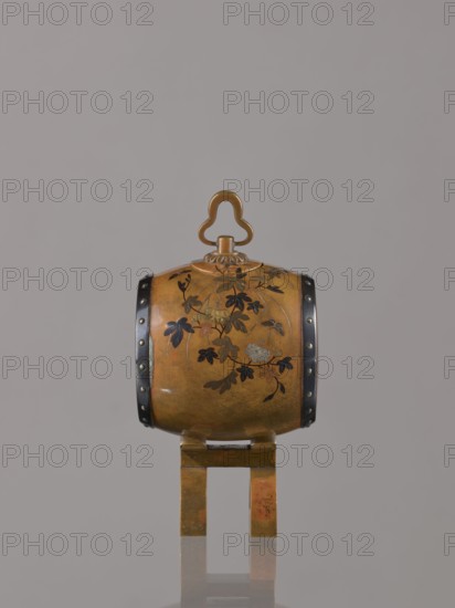 Okimono in the shape of a taiko drum, maker unknown Nogawa workshop ?, ca. 1890–1900 Meiji period, 1868–1912, Japan, Kyoto. Drum-shaped incense burner, carved/formed as a taiko drum; decorated with butterflies, insects, plant motifs including paulownia. Height 15.0 cm; diameter 7.8 cm., An ornate rounded object with a warm golden-brown surface sits on a matching golden-brown stand, decorated with a floral vine pattern in dark brown and black, with touches of lighter metallic tones; the object has two dark vertical bands with small round studs on either side and a clover-shaped handle at the top in a similar golden color, all set against a plain light gray background.