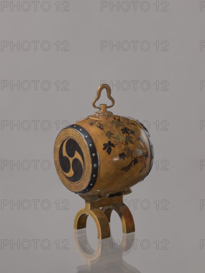 Okimono in the shape of a taiko drum, Nogawa workshop?, taiko drum, butterflies, insects, plant motifs, paulownia, Drum-shaped incense burner, ca. 1890–1900 Meiji period, 1868–1912, Japan, Kyoto, vessel; height 15.0 cm, dia. 7.8 cm, An ornate small drum-like object with a rounded body in golden brown tones sits on a matching golden brown stand with arched legs; the drum has decorative black painted floral motifs and leaf shapes on its surface, a circular end panel with a large black comma-like triple motif surrounded by a patterned border, dark metallic riveted bands at the drum edges, and a decorative golden brown looped handle on top; the background is plain light gray and the object casts a soft reflection on the surface below.