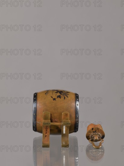 Okimono in the shape of a taiko drum, maker possibly Nogawa, ca. 1890–1900 Meiji period, 1868–1912. Drum-shaped incense burner, Japan, Kyoto., Japanese Meiji-period vessel with butterflies, insects, plant motifs including paulownia. Height 15.0 cm, diameter 7.8 cm., A small decorative drum and a separate drum beater sit on a reflective surface against a plain light gray background. The drum is round with a warm brown and amber body showing darker brown floral motifs and black metal bands with rivets around its edges; it rests on a simple brass-colored stand with a few vertical supports. The beater is similarly colored in brown and amber with darker floral decorations and a brass-colored handle ending in a rounded loop; both objects cast faint reflections on the surface. The visible colors include brown, amber, brass-colored metal, black, and light gray.