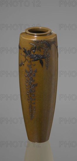 Vase, Nogawa role classification of creator, workshop; ; repossession Krakow — wisteria, butterflies. Meiji 1868–1912. A slender miniature vase with a belly widening slightly toward the top and narrow straight arms; no neck and a spout in the form of a flat ring. The bottom is round, slightly recessed, with an engraved brand. Decoration is sparse: a naturalistically treated wisteria vine placed in the upper part of the belly on one side, from which a sumptuous flower cluster hangs down almost to the end of the vase. The masterly technique of combining various metals and alloys gives the vessel colorfulness, enhanced by the darker tone of the decorative motif than the background. Although made of silver, the use of gilding and black shakudo patina makes it appear colorful, combining naturalism with sublime decorativeness. ca. 1890–1900 Meiji Period, Japan; Kyoto. Vessel; height 15.7 cm, width 5.3 cm., A tall, narrow vase with a rounded rim and a smooth, glossy surface in brown and amber...