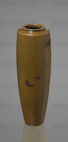A slender miniature silver vase by Nogawa, ca. 1890–1900 Meiji period, 1868–1912, Kyoto, Japan. The vase has a belly widening slightly toward the top, narrow straight arms, no neck, and a spout in the form of a flat ring; the bottom is round, slightly recessed, with an engraved brand. Decoration: a naturalistically treated wisteria vine placed on the upper part of the belly on one side, with a sumptuous flower cluster hanging down almost to the end. Made of silver with gilding and black shakudo patina, combining various metals and alloys to produce a darker decorative motif against the background, enhancing colorfulness and naturalistic yet decorative effect. Height 15.7 cm; width 5.3 cm., A tall cylindrical vase with a rounded rim, shown against a plain gray background, in a mottled brown and olive tone with a glossy finish; a few small dark metallic butterfly-shaped decorations and a small cluster of dark leaf-shaped accents are attached to its surface.