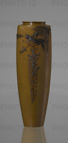 A slender miniature vase, with a belly widening slightly towards the top and narrow, straight arms. There is no neck, and the spout is in the form of a flat ring. The bottom is round, slightly recessed, with an engraved brand. The decoration is sparse, consisting of a naturalistically treated wisteria vine placed in the upper part of the belly on one side, from which a sumptuous flower cluster hangs down, almost to the end of the vase. Thanks to the masterly technique of combining various metals and alloys in one work, characteristic of Japanese craftsmanship of that period, the vessel gained colorfulness, enhanced by the darker tone of the decorative motif than the background. Although made of silver, through the use of gilding and black shakudo patina it is also colorful, enhancing the effect of fidelity to nature in the depiction of the plant, where naturalism is combined with sublime decorativeness. Maker: Nogawa. Role: creator/workshop. Date: ca. 1890–1900 Meiji Period, 1868–1912....