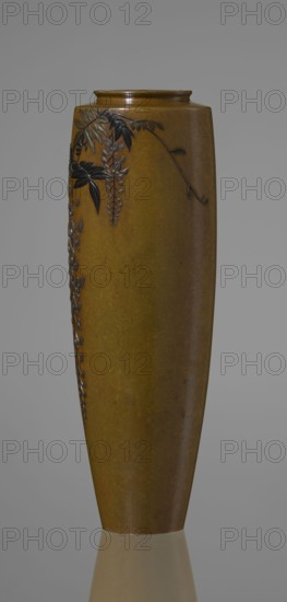 Vase, Nogawa role classification of creator, workshop; ; repossession Krakow — wisteria, butterflies; Japanese art, vases, Japanese culture; Meiji 1868–1912. A slender miniature vase with a belly widening slightly toward the top and narrow, straight arms. There is no neck; the spout is a flat ring. The bottom is round, slightly recessed, with an engraved brand. Decoration is sparse: a naturalistically treated wisteria vine on the upper part of the belly on one side, with a sumptuous flower cluster hanging almost to the base. Masterly technique of combining various metals and alloys gives the vessel colorfulness, enhanced by the darker tone of the decorative motif versus the background. Although made of silver, gilding and black shakudo patina make it appear colorful, combining naturalism with sublime decorativeness. Ca. 1890–1900 Meiji Period, Japan; Kyoto. Vessel. Height 15.7 cm; width 5.3 cm., A tall tapered vase with a smooth surface in shades of brown and ochre stands against a gra...