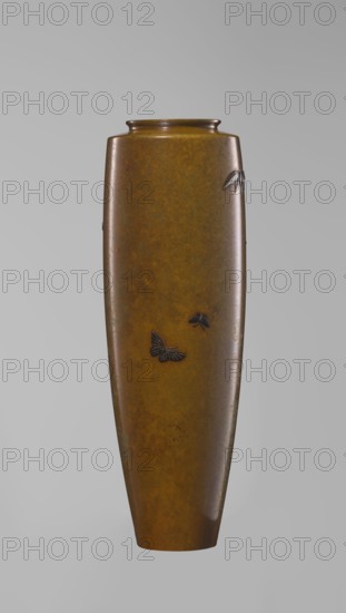 A slender miniature silver vase by Nogawa, ca. 1890–1900 Meiji period, 1868–1912, Kyoto, Japan. Belly slightly widening toward the top with narrow straight arms, no neck, spout formed as a flat ring; round, slightly recessed bottom with an engraved brand. Sparse decoration: a naturalistically treated wisteria vine on the upper belly with a sumptuous hanging flower cluster nearly reaching the base. Made using masterful combination of various metals and alloys, employing gilding and black shakudo patina so the silver vessel appears colorful, the darker toned motif enhancing naturalistic yet decorative depiction. Height 15.7 cm; width 5.3 cm., A tall, narrow vase with a smooth, slightly tapered cylindrical shape in mottled brown and golden tones stands against a plain light gray background; a few small dark decorative elements resembling butterflies and leaves are attached to its surface, and the vase has a subtle glossy finish with variations of darker and lighter brown throughout.