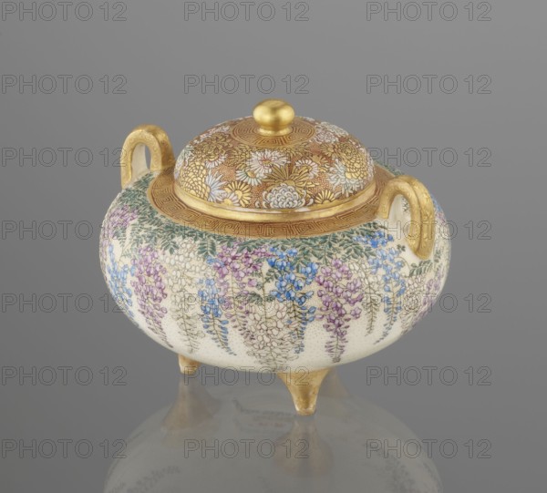 Incense burner koro with cover by Kinkozan Sobei VII 1867–1927 and Kinkozan workshop, signed. Meiji period 19th/20th century, Kyoto Awata, Japan. Ceramic satsuma censer koro, tripod with two handles, decorated with wisteria, chrysanthemums, and flowers; okimono style. Height 7.8 cm; diameter 9.5 cm., A rounded ceramic vessel with a domed lid and two loop handles sits on three short feet; the lid and rim are gilded and decorated with intricate floral motifs in gold and white, while the body is covered in a dense pattern of hanging floral clusters in shades of blue, purple, pink, green, and white against a pale cream background; the handles and feet are gilded, and the overall color palette includes gold, cream, blue, purple, pink, green, and white, with a neutral gray background and a subtle reflection beneath the object.