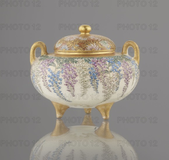Incense burner koro with cover, Kinkozan Sobei VII 1867–1927, Kinkozan workshop co-creator. Meiji period 19th/20th century, Kyoto Awata, Japan. Satsuma ceramics censer kyo-satsuma, okimono-style, decorated with wisteria and chrysanthemums; tripod form with two handles. Signed. Height 7.8 cm; diameter 9.5 cm., A rounded lidded vessel with a pair of loop handles and three short tapered legs sits against a neutral background; the lid and handles have a metallic gold finish and the lid top has a rounded gold knob, while the body is a creamy off-white decorated with hanging floral clusters in shades of blue, purple, and pale pink with green foliage, and the gold elements show a subtle engraved pattern; the vessel and its legs are reflected on a glossy surface below, and the overall color palette includes gold, cream, blue, purple, pink, and green.