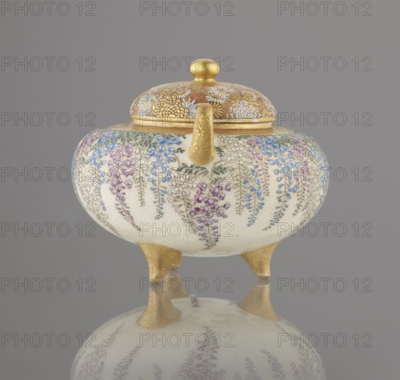 Incense burner koro with cover, Kinkozan Sobei VII 1867–1927, Kinkozan workshop co-creator. Meiji period 19th/20th century, Kyoto, Awata, Japan. Ceramics Kyoto satsuma, vessel on three legs with two handles; wisteria, chrysanthemums, and other flowers decoration; okimono-style censers. Signed. Height 7.8 cm; diameter 9.5 cm., An ornate rounded container with a domed lid sits on three short tripod legs, all parts showing a metallic gold finish on the lid knob, rim, spout, and legs; the body of the container is a creamy off-white decorated with cascading floral patterns in shades of purple, blue, green, and lilac, with delicate green leaves and fine dotted texture across the surface, set against a neutral gray background and reflected faintly below.