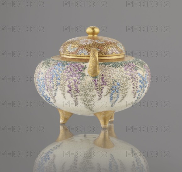 Incense burner koro with cover, Kinkozan Sobei VII 1867–1927 maker; workshop Kinkozan, signed. Meiji period 19th/20th century, Kyoto Awata, Japan. Ceramics: Kyoto satsuma; vessel with wisteria, chrysanthemums, flowers; on three legs and with two handles; tripod censers/okimono. Height 7.8 cm; diameter 9.5 cm., An ornate, rounded lidded vessel resting on three short feet, with a domed lid topped by a rounded knob and a short curved handle; the upper section and feet are metallic gold, and the main body is a creamy off-white decorated with cascading floral motifs in shades of purple, blue, green, and pale pink, with delicate fine-line detailing and small dotted texture; the lid also features intricate floral patterns in gold and pale tones; the object sits on a reflective surface that mirrors its colors and shapes against a neutral gray background.