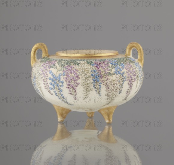Incense burner koro with cover by Kinkozan Sobei VII 1867–1927, with Kinkozan workshop co-creator. Ceramics Kyoto satsuma, Meiji period 19th/20th century. Satsuma censer decorated with wisteria, chrysanthemums and flowers; okimono-style, tripod form with two handles. Signed. Vessel: height 7.8 cm; diameter 9.5 cm. Origin: Awata, Kyoto, Japan., A rounded ceramic vessel with a pale cream background decorated with hanging floral motifs in shades of blue, purple, pink, white, and green stands on three short gold-colored feet and has two ornate gold-colored handles on opposite sides; the top rim is also gold-colored and the piece is reflected on a glossy gray surface against a plain gray background.