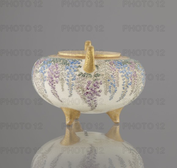 Incense burner koro with cover by Kinkozan Sobei VII 1867–1927, Kinkozan workshop; ceramics Kyoto satsuma, Meiji period 19th/20th century. Earthenware censers/koro; three-legged vessel with two handles, decorated with wisteria, chrysanthemums and flowers; signed. Height 7.8 cm; diameter 9.5 cm. Japan: Kyoto, Awata., A rounded ceramic vessel with a pale cream background sits on three short gold-colored legs and has a gold-colored rim and handle; the body is decorated all around with hanging clusters of small flowers in shades of purple, blue, and pink with green leaves, and the surface shows a fine dotted texture; the gold-colored legs and handle have subtle floral embossing, and the vessel and its legs are reflected on a smooth gray surface beneath; the background is a uniform muted gray.