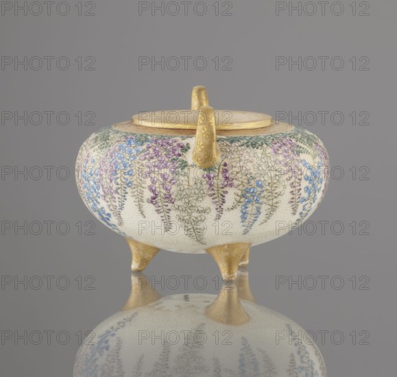 Incense burner kôro with cover by Kinkôzan, Sôbei VII 1867–1927, co-created by Kinkôzan workshop; ceramics Kyoto Satsuma, Meiji period 19th/20th century, Japan, Kyoto, Awata. Satsuma censer on three legs with two handles, decorated with wisteria, chrysanthemums and flowers; okimono-style; signed. Height 7.8 cm; diameter 9.5 cm., A rounded ceramic vessel with a lid and three short legs sits against a gray background, decorated with hanging floral motifs in shades of purple, blue, green, and white on a cream surface, with gold-colored handles, rim, and legs; the vessel and its decoration are reflected on the glossy surface below, and the overall scene includes tones of gray, cream, gold, purple, blue, and green.