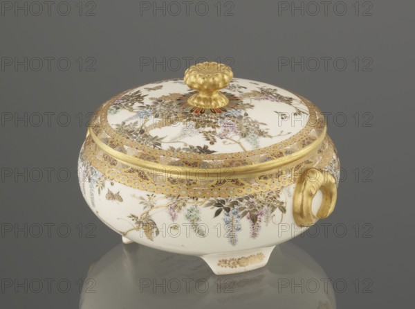 Small box with a lid, Kinkôzan, Sôbei VII 1867–1927 maker, Kinkôzan workshop, Japanese, Meiji period late 19th century, Kyoto, Awata. Ceramic satsuma box decorated with wisteria and butterflies. The lid has a gold knob shaped like a flower; the vessel has two slightly convex handles and stands on three wide feet. Signed. Height 6.8 cm; diameter 10.2 cm., A lidded round ceramic bowl with a domed lid and a decorative knob sits on a reflective surface; the bowl is predominantly white with extensive gold ornamental trim and a gold knob, and it features painted floral and foliage motifs in colors including green, blue, pink, purple, and brown; a gold handle is visible on one side and the piece rests on small feet with gold detailing, all set against a uniform grey background.