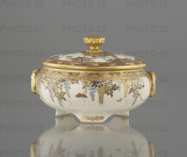 Small box with a lid, Kinkôzan, Sôbei VII 1867–1927 author, Kinkôzan workshop / co-creator; Japanese, Meiji late 19th century; Kyoto, Awata. Satsuma ceramics vessel decorated with wisteria and butterflies. The lid has a gold knob in the shape of a flower. The vessel has two slightly convex handles on three wide feet. Signed. Height 6.8 cm; diameter 10.2 cm., A small lidded porcelain bowl with a rounded body and short feet sits on a reflective surface against a gray background; the bowl is primarily white with ornate gold trim and gilded handles on each side, and the lid has a gold knop; delicate painted floral garlands in colors including blue, pink, green, purple, and brown hang around the body and lid, with small painted insects in red and yellow visible among the flowers; the reflection of the bowl and its decoration is visible on the surface below.