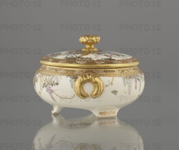 Small box with a lid, Kinkôzan, Sôbei VII 1867–1927 author, Kinkôzan workshop/co-creator; ; Japanese art; Japanese; Meiji 1868–1912; wisteria; ceramics; butterflies; boxes; satsuma. The lid with a gold knob in the shape of a flower. The vessel with two slightly convex handles, on three wide feet. Signed, late 19th century Meiji period. Japan; Kyoto, Awata. Vessel, container; height 6.8 cm; diameter 10.2 cm., A small lidded ceramic vessel with a rounded white body and a matching domed lid, both decorated with delicate painted floral motifs in muted purples, greens, and pale blues, and detailed gold-painted borders and accents; the lid has a textured gold knob, and the vessel's body features a prominent gold ornamental handle on the front and several small gold decorative trims, all resting on short feet and reflected on a smooth gray surface, with the background a uniform gray.
