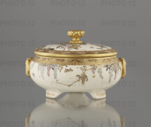 Small box with a lid. Maker: Kinkôzan Sôbei VII 1867–1927 and workshop Kinkôzan. Satsuma ceramics, late 19th century Meiji period, Kyoto, Awata, Japan. The lid has a gold knob in the shape of a flower. The vessel has two slightly convex handles, stands on three wide feet, and is decorated with wisteria and butterflies. Signed. Height 6.8 cm; diameter 10.2 cm., An ornate lidded ceramic dish with a rounded body and short feet sits against a plain gray background, showing a glossy reflection beneath. The lid has a rounded golden knob and a broad gold rim with intricate decorative patterns, while the body is primarily off-white with delicate painted floral garlands and hanging clusters in pastel shades including pink, lavender, blue, and green, accented with gold detailing. There are gold handles on each side and gold ornamentation along the upper edge and feet, with small painted botanical motifs and an overall polished, finely decorated appearance.