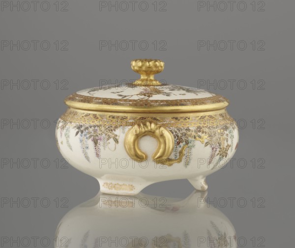 Small box with a lid, Kinkôzan, Sôbei VII 1867–1927 author, Kinkôzan workshop, Japanese, Meiji late 19th century, Japan, Kyoto, Awata. Satsuma ceramics vessel decorated with wisteria and butterflies. The lid has a gold knob in the shape of a flower. The vessel has two slightly convex handles and stands on three wide feet. Signed. Height 6.8 cm; diameter 10.2 cm., A small lidded ceramic vessel with a rounded body and short feet sits against a gray background, the piece predominantly white with extensive gold ornamentation on the lid, rim, handle, and decorative elements; delicate painted floral motifs in shades of purple, blue, pink, green, and brown cascade around the body beneath a band of intricate gold patterning, and a gold knob tops the lid while a gold ornamental handle-like feature decorates the front, with the vessel and its colors reflected faintly on the glossy surface below.