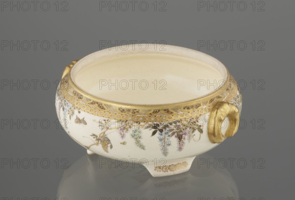 Small box with a lid, Kinkôzan Sôbei VII 1867–1927 maker; Kinkôzan workshop. Satsuma ceramics, late 19th century Meiji period, Japan — Kyoto, Awata. Wisteria and butterflies decoration. Lid with a gold knob shaped like a flower; vessel with two slightly convex handles on three wide feet. Signed. Height 6.8 cm; diameter 10.2 cm., A low, round ceramic bowl with a pale cream interior and an off-white exterior sits on a reflective gray surface against a gray background. The bowl has a decorative gold rim and a band of ornate gilded patterning below the rim. Floral garlands in soft colors including pink, blue, green, and purple hang around the exterior among small painted leaves in brown and green. There are gilded handles on the sides with a curved, sculpted shape and a few small gilded decorative accents on the short feet. The overall visible colors include cream, off-white, gold, gray, pink, blue, green, purple, and brown.