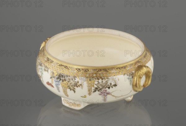 Small box with a lid, Kinkôzan, Sôbei VII 1867–1927 maker, Kinkôzan workshop, Japanese, Meiji period late 19th century. Satsuma ceramics decorated with wisteria and butterflies. The lid has a gold knob shaped like a flower. The vessel has two slightly convex handles and stands on three wide feet. Signed. Vessel/container; height 6.8 cm, diameter 10.2 cm. Origin: Awata, Kyoto, Japan., A small oval ceramic bowl with a creamy off-white interior and exterior sits on a reflective gray surface, decorated around its upper rim with an ornate gold band and delicate painted floral motifs in shades of gold, green, blue, pink, and brown, and featuring a gold-colored loop handle on one side and short molded feet at the base; the overall visible colors include cream, gold, gray, green, blue, pink, brown, and a hint of red.