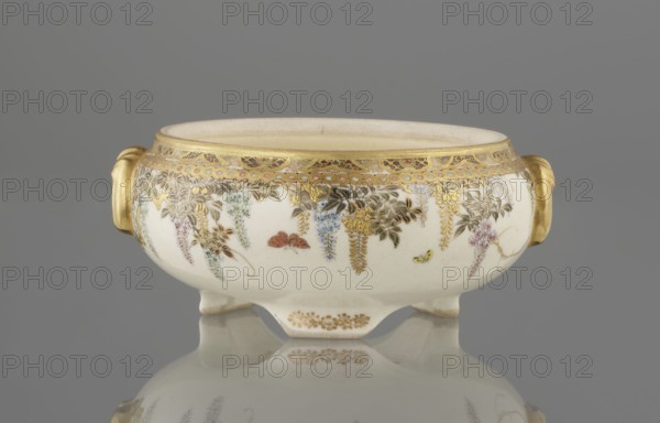 Small box with a lid, Kinkôzan, Sôbei VII 1867–1927 maker, Kinkôzan workshop, Japanese, Meiji period late 19th century. Satsuma ceramics decorated with wisteria and butterflies. The lid has a gold knob in the shape of a flower. The vessel has two slightly convex handles and stands on three wide feet. Signed. Kyoto, Awata. Height 6.8 cm; diameter 10.2 cm., A low, rounded ceramic bowl with a pale cream base color sits on a reflective surface, showing a mirrored reflection beneath; the bowl has a wide rim with a gilded band and ornate gold decorative border, and gold handles on each side. Along the exterior there are delicate painted hanging floral motifs in shades of purple, blue, pink, green, and brown, with additional gold leaf accents and small painted insects in red and yellow. The bowl rests on short feet that are also gilded, and the background behind the bowl is a uniform gray.
