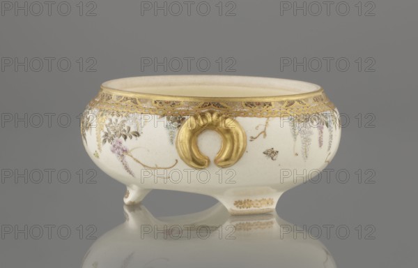 Small box with a lid, Kinkôzan, Sôbei VII 1867–1927 maker; Kinkôzan workshop. . Japanese, Meiji period late 19th century. Ceramics satsuma. Kyoto, Awata. The lid has a gold knob in the shape of a flower. The vessel has two slightly convex handles and rests on three wide feet. Signed. Subject motifs: wisteria and butterflies. Vessel/container. Height 6.8 cm; diameter 10.2 cm., A low, rounded ceramic bowl with a glossy white surface sits on small feet and casts a reflection on a smooth gray surface, decorated with gold ornamental trim around the rim and a prominent raised gold handle-like motif on the side, with delicate painted floral garlands and hanging blossoms in soft purple and pale blue tones accented by fine brown stems and green leaves, and small touches of brown in tiny insect-like details against a neutral beige interior and a muted gray background.