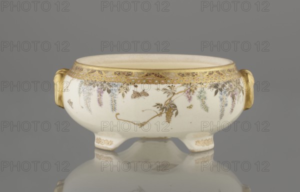 Small box with a lid, Kinkôzan, Sôbei VII 1867–1927 creator / craftsman; Kinkôzan workshop / co-creator; Japanese, Meiji late 19th century, 1868–1912; Kyoto, Awata. Satsuma ceramics vessel/container with wisteria and butterflies decoration. The lid has a gold knob in the shape of a flower. The vessel has two slightly convex handles and stands on three wide feet. Signed. Height 6.8 cm; diameter 10.2 cm., A low, round ceramic bowl with a cream base color sits on a reflective surface against a gray background; it has a decorative gold rim and gold handles on each side, small tapered feet with gold ornamentation, and a painted floral garland of hanging wisteria-like clusters in shades of purple, blue, pink, and pale green interspersed with gold leaves and a few small painted insects in brown and yellow.