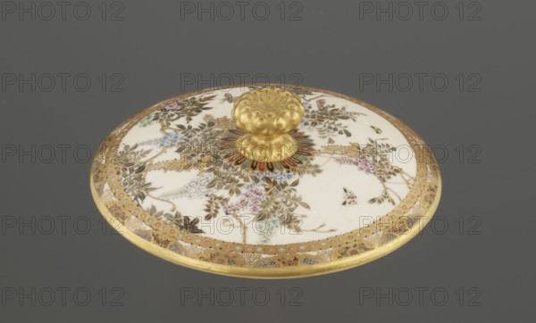 Small box with a lid, Kinkôzan, Sôbei VII 1867-1927 author; Kinkôzan workshop/co-creator; wisteria, ceramics, butterflies, boxes, satsuma. The lid with a gold knob in the shape of a flower. The vessel with two slightly convex handles, on three wide feet. Signed, late 19th century Meiji period, Japan, Kyoto, Awata. Vessel/container; height 6.8 cm, diameter 10.2 cm., A round decorative lid with a central raised knob sits against a gray background; the knob and outer rim are gold, and the flat surface is primarily white with ornate floral and foliate patterns in gold, green, blue, pink, and brown, plus a few small butterfly-like motifs; a colorful radial motif in red, green, and blue surrounds the base of the knob, and a gold patterned border runs around the edge.