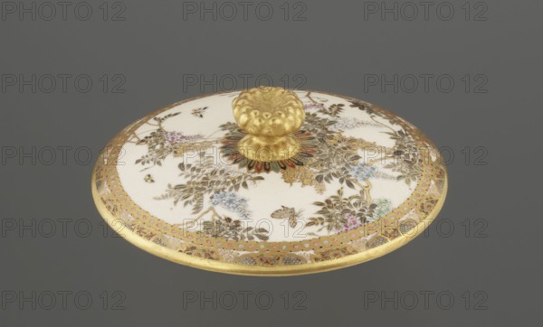 Small box with a lid, Kinkôzan, Sôbei VII 1867–1927 maker, Kinkôzan workshop, Japanese, Meiji period late 19th century, Japan; Kyoto, Awata. Satsuma ceramic vessel with wisteria and butterflies decoration. The lid has a gold knob shaped like a flower. The vessel has two slightly convex handles and stands on three wide feet. Signed. Height 6.8 cm; diameter 10.2 cm., A round decorative lid with a central raised knob sits against a grey background; the lid has a white surface decorated with floral and leafy patterns in shades of green, brown, blue, pink, and purple, with a gold rim and gold detailing throughout, and the central knob and surrounding medallion are gold with accents of red, blue, and green.