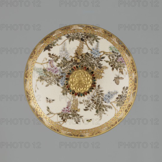 Small box with a lid, Kinkôzan, Sôbei VII 1867–1927 author; Kinkôzan workshop; . Japanese, Meiji period late 19th century. Satsuma ceramic vessel decorated with wisteria and butterflies. The lid has a gold knob shaped like a flower. The vessel has two slightly convex handles and stands on three wide feet. Signed. Origin: Kyoto, Awata. Height 6.8 cm; diameter 10.2 cm., A circular decorative plate shown against a gray background, featuring a central raised gold medallion surrounded by a radiating floral motif with accents of red, green, and dark blue; the plate surface is primarily creamy white with painted botanical designs in shades of brown, green, pale blue, pink, and lavender, and includes several small insects depicted in brown and black; an ornate gold border with intricate patterns encircles the rim, and additional gold detailing appears throughout the foliage and floral elements.