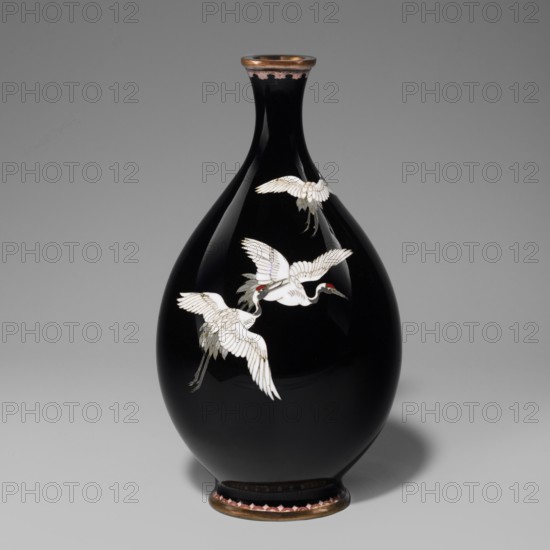 Vase, cloisonné enamel, Meiji period 19th/20th century, 1868–1912, attributed to Nagoya, Japan. Cylindrical vessel with a thin short neck widening toward the spout; decorated with cranes iconography. Height 18.3 cm; diameter 10 cm.,  noted., A teardrop-shaped glossy black vase with a narrow neck and flared rim sits against a gray background, decorated with a group of white birds in flight with touches of red and pale gray on their heads and wings, and gold accents outlining some feathers; the rim and foot of the vase have a metallic bronze tone with a band of pink and darker scalloped detailing, and the vase casts a soft shadow on the surface beneath it.