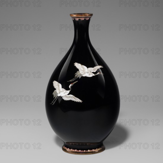 Vase, cloisonné enamel, Meiji period 1868–1912, 19th/20th century. Nagoya, Japan. Vessel with a thin short neck widening towards the spout; decorated with cranes iconography. Height 18.3 cm; diameter 10 cm., A glossy black teardrop-shaped vase with a narrow neck and flared rim, decorated with a pair of white cranes in flight with touches of gray and red on their heads and beaks; the rim and foot have metallic bronze tones with a band of pinkish accents along the base, and the background is a smooth gray gradient.