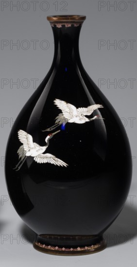 Vase, gift; cloisonné enamel; Japanese; Meiji period 1868–1912, 19th/20th century; Nagoya, Japan; subject: cranes iconography; With a thin, short neck widening towards the spout; vessel; height 18.3 cm, diameter 10 cm., A glossy black vase with a rounded body and a narrow neck, featuring a metallic rim at the top and a metallic base trim at the bottom; the body shows a decorative motif of a few white cranes in flight with pale gray and muted green tail feathers and small red accents on their heads, and there are subtle reflections and highlights on the vase surface showing deep black and dark gray tones against a neutral gray background.