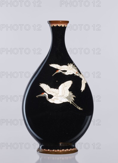 Vase, Meiji period 1868–1912, late 19th/early 20th century; Japanese cloisonné enamel; Nagoya, Japan; decorative vessel vase depicting birds—cranes; height 18.3 cm, diameter 10 cm., A teardrop-shaped vase with a glossy black surface decorated with a pair of white cranes in flight, each crane showing white feathers with touches of gray and small red markings on their heads; the vase has a narrow neck topped with a brown rim and a decorative band below the rim in shades of beige and red, and a matching brown base with a scalloped beige and red border; the background is a pale light gray.