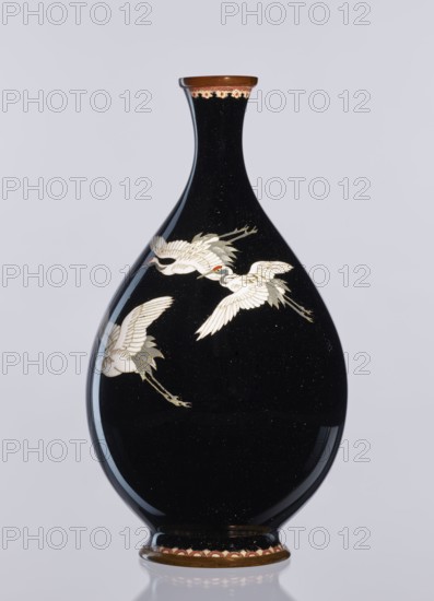 Vase, Meiji period 1868–1912, late 19th/early 20th century; Japanese cloisonné enamel; Nagoya, Japan; decorative vessel/vase height 18.3 cm, diameter 10 cm; decoration: birds—cranes iconography., A glossy black, teardrop-shaped vase with a narrow neck and flared rim sits against a pale gray background, decorated with a few white cranes in flight whose feathers show subtle gray and gold accents and small red markings near their heads; the rim and base have brown and gold bands with a scalloped decorative border in muted red and cream, and the vase surface reflects light creating highlights and a few tiny speckles.