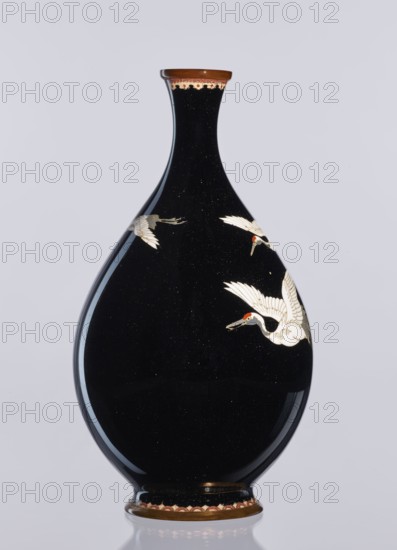 Vase, Meiji period 1868–1912, Nagoya, Japan; cloisonné enamel vessel decorated with birds/cranes; height 18.3 cm, diameter 10 cm; 19th/20th century., A glossy black teardrop-shaped vase with a narrow neck and flared rim, decorated with several white birds in flight each showing small red accents on their heads, a brown rim at the top with a patterned band in light and dark tones, and a matching brown base with a decorative scalloped band, all shown against a pale gray background.