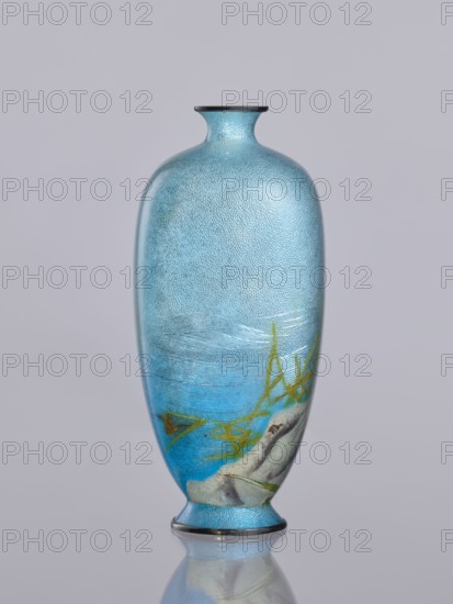 Vase, Tomiki, Shôbei fl. ca. 19th/20th century, chidori plovers, reeds, waves; cloisonné enamel; Meiji period 1868–1912, Japan; decorative vessel vase; signature: Tomiki engraved on the bottom in a vase-shaped cartouche — 3 katakana characters: Tomiki Tomiki Sobei; height 12.2 cm, width 5.6 cm, depth 5.6 cm., A tall, narrow vase with a rounded body and a short flared neck stands against a light gray background, its surface showing a textured, speckled blue that ranges from pale to medium tones. Near the lower portion the blue deepens into a richer shade around a painted scene that includes a light gray rock-like shape and a group of thin, angular green plant forms. The vase has a thin dark rim at the top and a matching dark band at the small foot, and the base and lower area reflect faintly on the smooth surface below.