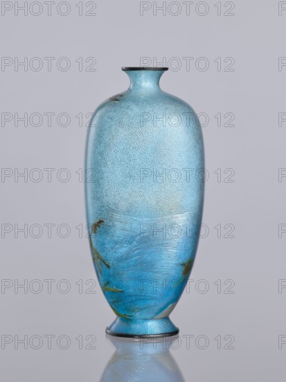 Vase, Tomiki, Shôbei fl. ca. 19th/20th century craftsman, chidori plovers, reeds, waves; cloisonné enamel; Meiji period 1868–1912, Japan; decorative vessel; Signature: Tomiki engraved on the bottom in a vase-shaped cartouche with 3 katakana characters: Tomiki Tomiki Sobei, 19th/20th century; height 12.2 cm, width 5.6 cm, depth 5.6 cm., A tall, slender vase with a narrow neck and flared rim sits centered against a pale gray background, its surface showing a textured, pebbled finish in shades of light blue and turquoise that deepen toward the lower half, where sweeping horizontal brushlike strokes create a band of richer blue with hints of green. The vase has subtle darker accents at the rim and base in a deep gray or black, and there are a few delicate, thin, plantlike motifs in muted green and brown near the lower portion. The vase rests on a small foot and casts a soft reflection on the glossy surface beneath it.
