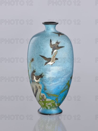 Vase, Tomiki, Shôbei fl. ca. 19th/20th century craftsman, chidori plovers, cloisonné enamel, Japanese art, reeds, waves, decorative vessel, Meiji 1868–1912, Signature: Tomiki engraved on the bottom in a vase-shaped cartouche, 3 katakana characters: Tomiki; artist Tomiki Sobei, 19th/20th century Meiji period, Japan; vessel; height 12.2 cm, width 5.6 cm, depth 5.6 cm, A tall, rounded vase with a narrow neck and flared rim sits against a pale gray background; the vase is predominantly light blue with a textured, speckled surface and areas of deeper blue, green, brown, white, black, and gray. The body of the vase is decorated with a scene of several birds in various flying and perched poses, their feathers rendered in shades of brown, white, black, and gray with small red accents on their beaks or feet. Near the base and lower sides there are stylized green leaves and stems, and a rocky motif in gray and brown. Subtle wave-like markings in lighter blue and white suggest water across the mi...