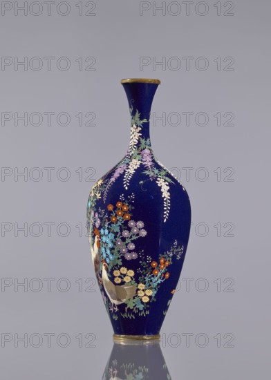 Vase, cloisonné enamel, ovoid with elongated body and long thin neck slightly flared at the spout; navy blue background decorated with wisteria, hens and roosters, and flowers. Japanese, Meiji period c. 1890–1910., Vessel; height 15.6 cm, diameter 5.6 cm, width 6.4 cm., A tall, slender vase with a deep blue background and a narrow flared neck, decorated with a colorful floral pattern and a pair of birds; the flowers appear in shades of purple, lavender, orange, pale yellow, light blue, white, and green, with trailing white wisteria-like clusters, and the birds show brown, cream, and red tones; the rim and base have metallic gold bands, and the vase sits on a reflective surface that mirrors its lower portion against a plain light gray backdrop.