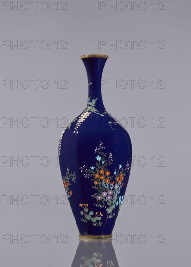 Vase, cloisonné enamel, wisteria, hens, flowers, roosters iconography, Japanese art, vases, Japanese culture, Meiji 1868–1912. Ovoid vase, elongated, with a long, thin neck, slightly flared at the spout. Navy blue background of the decoration. c. 1890–1910 Meiji period, Japan. Vessel; height 15.6 cm, dia. 5.6 cm, width 6.4 cm., A tall, narrow-necked vase with a deep blue background and a thin metallic rim at the top and base, decorated with several clusters of small floral motifs in colors including orange, light blue, pale purple, white, and green, with leafy green stems and small white accents, shown against a plain light gray background and reflected faintly on the surface below.