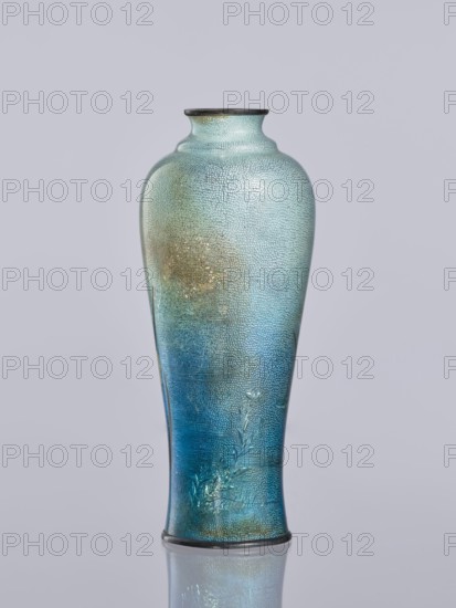 Vase by Miwa Tomisaburô fl. ca. late 19th–early 20th century. Cloisonné enamel elongated vase, Meiji period 1868–1912, early 20th century, Japan. Decoration: white and blue irises in the background; cranes engraved and polished in foil among plants, standing in water; foil covered with transparent enamel. Vessel. Height 12.2 cm; diameter 5.1 cm., A tall, narrow vase with a rounded shoulder and a slightly flared neck sits against a pale gray background, its surface showing a textured, pebbled finish and a subtle reflective sheen; the color transitions from pale aqua and light turquoise near the top through greenish-gold highlights to deeper teal and rich blue toward the lower half, and there is an embossed botanical motif near the base.