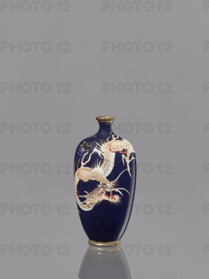 Vase, Japanese, Meiji 1868–1912, cloisonné enamel, 19th/20th century, depicts Japanese dragon motif/iconography, vessel, height 36.7 cm, diameter 4.6 cm., A dark blue ceramic vase with a narrow neck and flared rim sits upright against a gray background, featuring a decorative dragon motif in shades of cream, beige, red, and gold that winds around its body; the rim and base have gold-colored accents, and the vase casts a subtle reflection on the surface below.