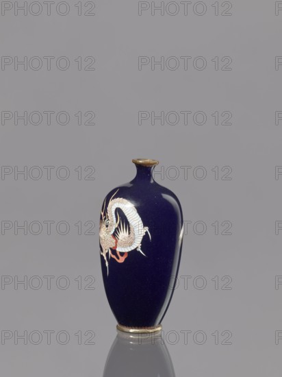 Vase, dragons iconography, Japanese art, Japanese dragon motif, Meiji 1868–1912, cloisonné enamel, 19th/20th century Meiji Period, Japan, vessel; height 9.5 cm, diameter 4.6 cm, A dark blue vase with a narrow neck and rounded body stands against a plain gray background; the vase features a decorative pale dragon-like creature with light tones and touches of red on one side, and the rim and base have metallic gold accents, with the vase casting a subtle reflection on the surface below.