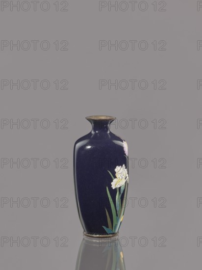 Vase, Ovoid cloisonné enamel vase with a short, thin, flared neck, dark blue background decorated with irises. Japanese, Meiji period ca. 1900. Height 9.1 cm; diameter 4.3 cm., A single tall vase sits upright against a plain gray background, the vase having a deep navy blue glossy surface with a narrow flared rim and a rounded body tapering slightly toward the base, decorated on one side with painted floral motifs including white and pale pink flowers and long green leaves, with subtle reflections visible on the vase and a faint mirrored reflection of the vase on the surface beneath it; visible colors include navy blue, white, pale pink, green, gray, and small areas of metallic brown around the rim and base.