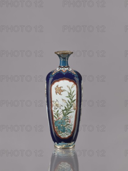 Vase, gift, revindication noted. Ovoid, elongated cloisonné enamel vase with a short, thin, strongly flared neck. Decoration: four reserves depicting blossoming cherry, apple tree, wisteria, and lilies on a navy blue background. Japanese, ca. 1910–1920 Meiji/Taisho periods. Vessel. Height 15.5 cm; diameter 6.0 cm., A tall, slender vase with a narrow flared neck and rounded shoulders sits against a plain gray background, its surface primarily deep blue with panels of white on the front and sides containing painted floral scenes. The floral panels show green leaves and stems with flowers in shades of pale yellow, white, and hints of pink, and there are teal or turquoise leaves near the base; the panels are bordered by a reddish-brown outline. The neck and shoulder area feature decorative motifs in blue, turquoise, gold, and white, and the rim and base have metallic gold or brass-colored accents. The vase casts a soft reflective image below, mirroring its colors and shapes on a glossy sur...