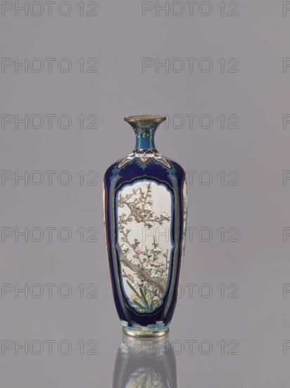 Vase, gift, revindication; ovoid elongated cloisonné enamel vase with a short, thin, strongly flared neck. Decoration: four reserves depicting blossoming cherry, apple tree, wisteria, and lilies on a navy blue background. Japanese, ca. 1910–1920 Meiji/Taisho periods. Vessel. Height 15.5 cm; diameter 6.0 cm., A tall, narrow vase with a flared rim standing against a plain gray background, predominantly deep blue with panels of off-white on its sides showing a painted branch with blossoms in pale pink and white and green leaves, decorative accents in shades of blue, teal, gold, and brown around the neck and base, and a reflective surface beneath the vase showing a muted reflection.