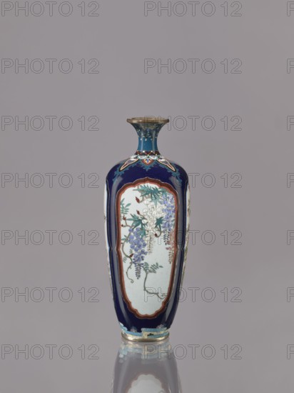 Vase, Ovoid, elongated cloisonné enamel vase with a short, thin, strongly flared neck. Decoration: four reserves depicting blossoming cherry, apple tree, wisteria, and lilies on a navy blue background. Subject: flowers iconography; style: Japanese art. Date: ca. 1910–1920 Meiji/Taisho periods. Material: cloisonné enamel. Function: vessel. Dimensions: height 15.5 cm; diameter 6.0 cm., A tall, narrow vase with a flared rim and a rounded shoulder, shown standing on a reflective surface; the vase has a deep blue background with panels of pale off-white featuring painted floral clusters and trailing vines in shades of blue, purple, green, and white, framed by reddish-brown borders; the neck and rim area display decorative motifs in blue and gold tones, and the base has bands and scalloped patterns in light blue, cream, and gold; the overall color palette includes deep blue, pale off-white, blue, purple, green, reddish-brown, cream, and gold.