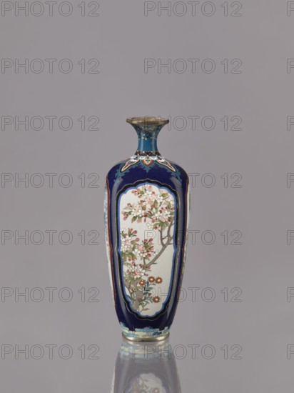 Vase, Ovoid elongated cloisonné enamel vase with a short, thin, strongly flared neck. Decoration: four reserves depicting blossoming cherry, apple tree, wisteria, and lilies on a navy blue background. Japanese; Meiji–Taisho period, ca. 1910–1920. Vessel; height 15.5 cm, diameter 6.0 cm., A tall, narrow vase with a flared neck sits against a plain gray background, predominantly dark blue with panels of off-white featuring floral branch motifs in shades of pink, red, green, brown, and white, with gold and light blue decorative borders and accents, and a metallic rim and base showing reflections on the surface below.
