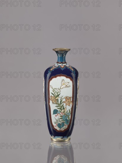 Vase, ovoid elongated cloisonné enamel vase with short, thin, strongly flared neck. Decoration: four reserves depicting blossoming cherry, apple tree, wisteria, and lilies on a navy blue background. Materials: cloisonné enamel. Date: ca. 1910–1920 Meiji–Taisho periods. Japan. Vessel. Height 15.5 cm; diameter 6.0 cm., A tall narrow vase with a flared lip, displaying a deep blue background and a central pale panel on its front and sides that contains painted floral motifs including lily-like flowers and leaves in colors such as white, yellow, green, teal, pink, and brown; the vase has decorative accents in gold and lighter blues around the neck and base, and the surface reflects faintly on the grayish background below.