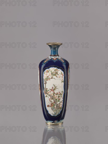 Vase, cloisonné enamel ovoid elongated vase with a short, thin, strongly flared neck. Decoration: four reserves depicting blossoming cherry, apple tree, wisteria and lilies on a navy blue background. Japanese, ca. 1910–1920 Meiji/Taisho periods. Vessel, height 15.5 cm, diameter 6.0 cm., A tall, narrow vase with a flared rim sits against a plain light gray background, the body of the vase predominantly deep blue with panels of off-white that display painted branches with blossoms and leaves in shades of pink, red, green, and brown, and small accents of gold outlining decorative borders; the neck of the vase is blue with turquoise and gold details, and the base shows bands of turquoise, gold, and blue with the vase casting a soft reflection on the glossy surface beneath it.